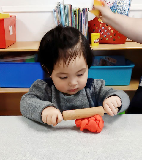 A focused child rolls playdough with a small rolling pin, engaged in a creative activity at a table surrounded by supplies.